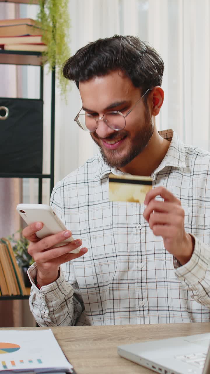 Felice uomo d'affari indiano che utilizza una carta di credito bancaria e uno smartphone per i pagamenti online.