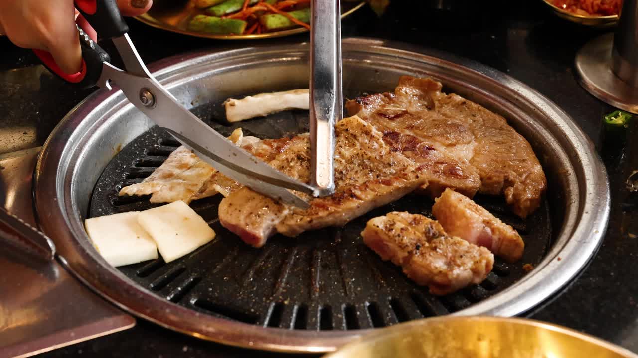 Hand uses scissors to cut grilled pork belly on tabletop Korean barbecue grill, warm lighting