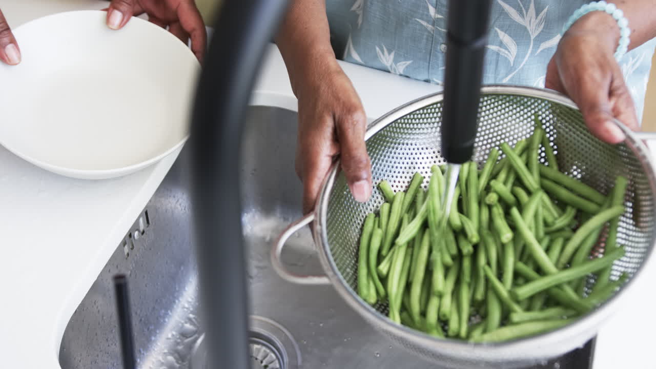 sección media de las amigas afroamericanas mayores lavando frijoles en el fregadero de la cocina, cámara lenta