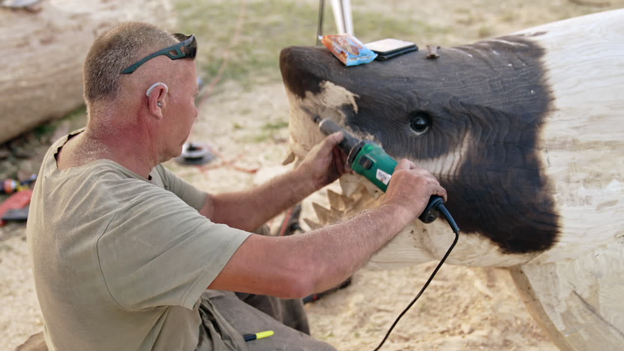 Male artist cutting a sculpture of shark from the wood. Artist applies electric tool in the process.
