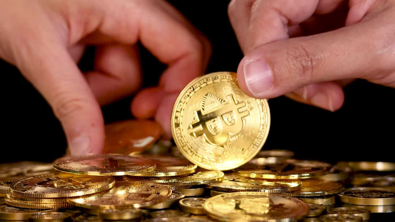 Human hand picks up shiny golden cryptocurrency coin from a pile under dramatic studio lighting