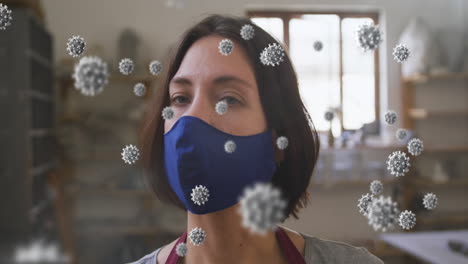 animation of covid 19 cells over woman in pottery studio wearing face mask