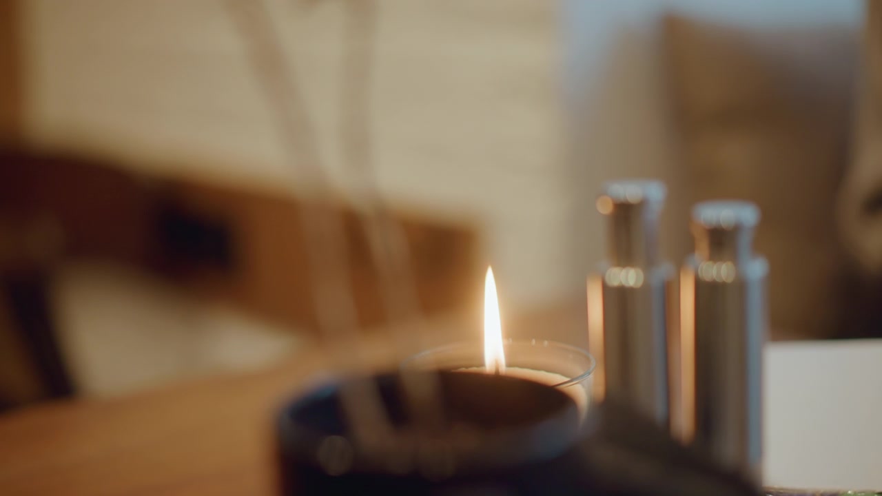 Gros plan sur une bougie allumée sur une surface en bois, avec une lumière douce et chaude et un arrière-plan flou. La flamme de la bougie vacille doucement, tandis que des objets métalliques se trouvent à proximité, créant une atmosphère intérieure calme.