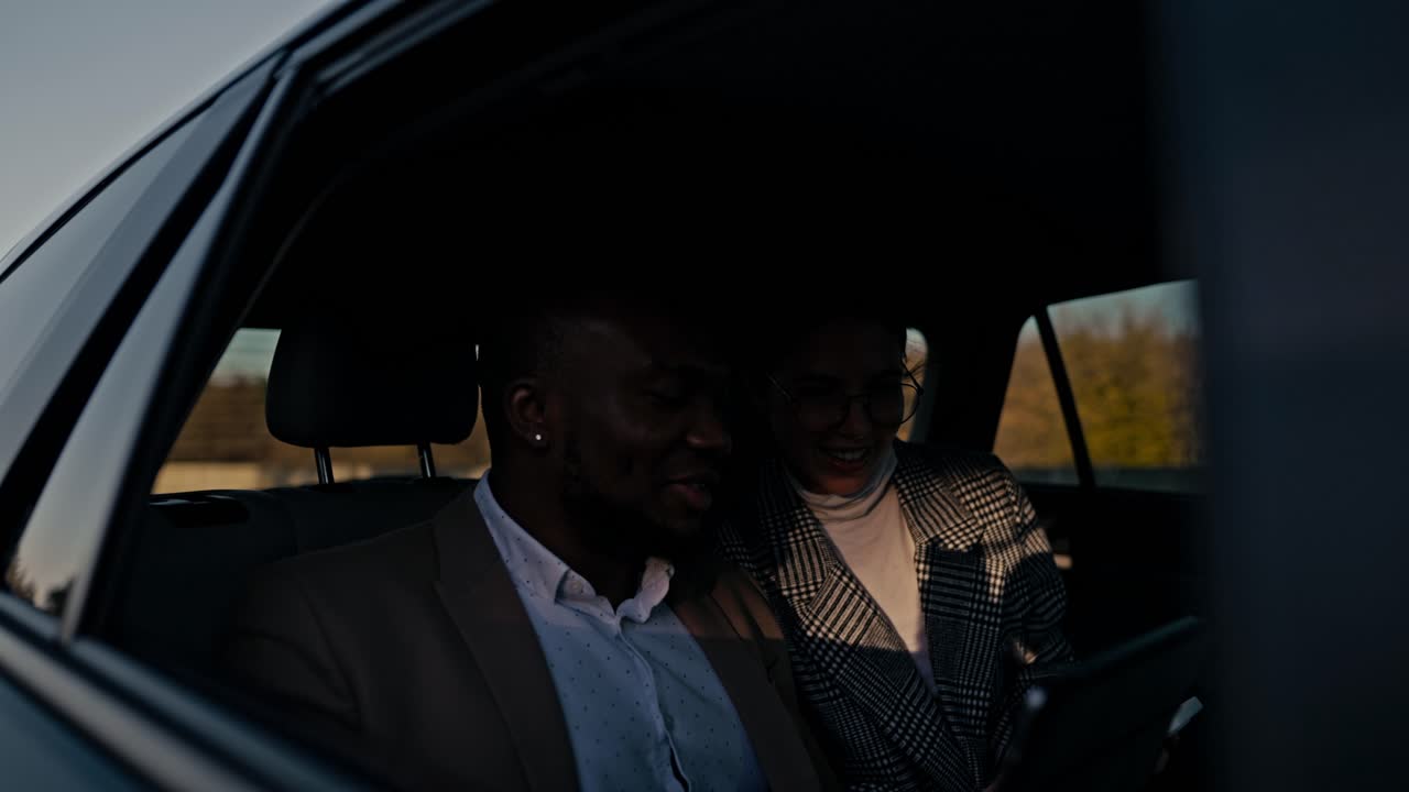 A happy man with black skin in a brown suit explains to a brunette girl in round glasses to his colleague about his thoughts and ideas using a tablet during a shared trip in a modern car