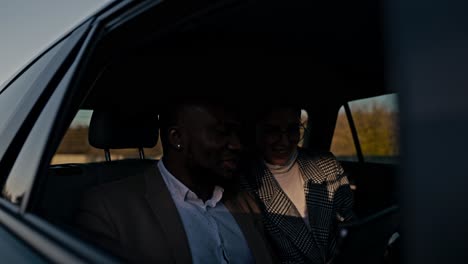 a happy man with black skin in a brown suit explains to a brunette girl in round glasses to his colleague about his thoughts and ideas using a tablet during a shared trip in a modern car