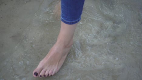 Female-bare-foot-standing-and-moving-sea-water-on-background-sandy-bottom