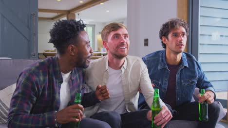 multi-cultural group of male friends watching sports game on tv at home celebrating goal with beer