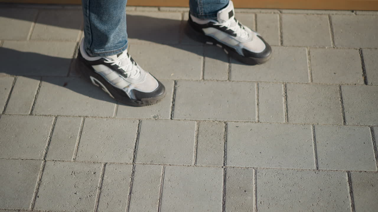 top down view of legs in jeans and white black sneakers walking slowly toward wooden bench on sunny day with long shadows falling on clean gray pavement in urban outdoor environment