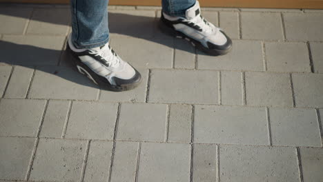 top down view of legs in jeans and white black sneakers walking slowly toward wooden bench on sunny day with long shadows falling on clean gray pavement in urban outdoor environment