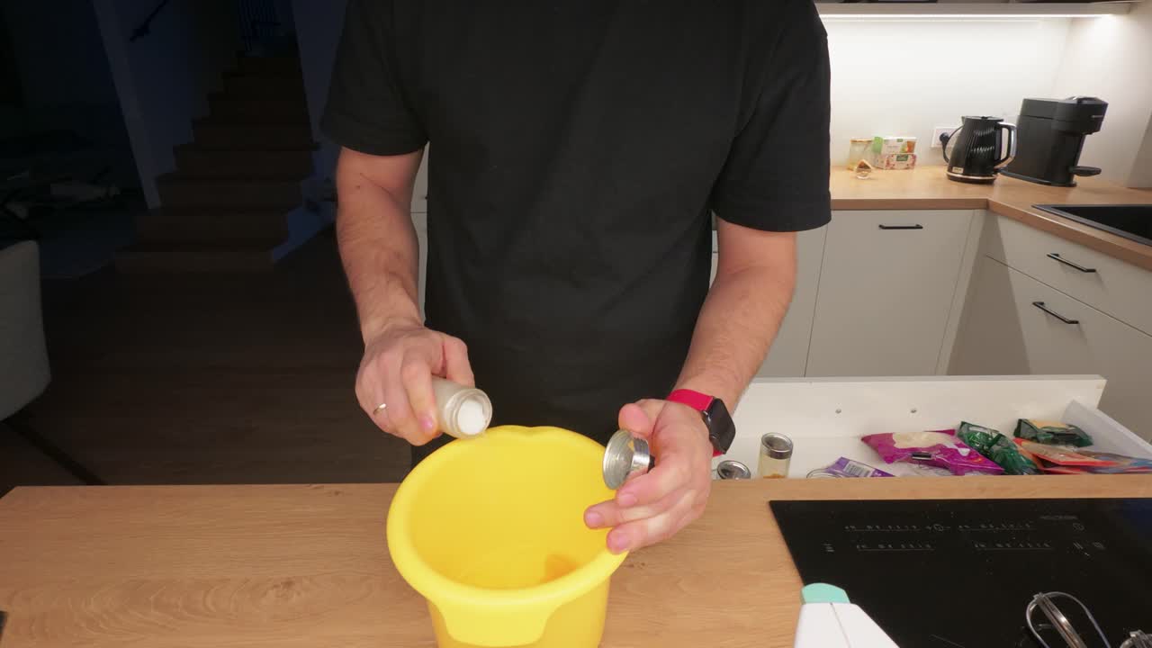 Inquadratura ravvicinata dettagliata di un uomo che prepara ingredienti in una cucina moderna. La cucina ben illuminata presenta un ampio piano di lavoro con spezie, utensili ed elettrodomestici visibili.