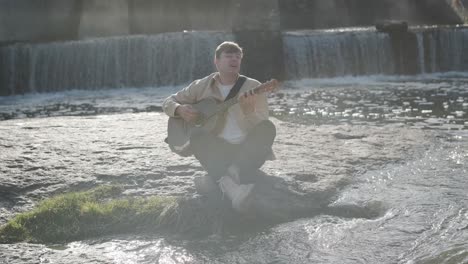 joven tocando la guitarra sentado en la orilla de un río de montaña sobre un fondo de rocas. concepto de libertad lugar de relajación.