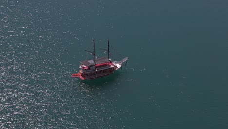 Vista-Aérea-Sobre-Un-Velero-De-Cubierta-Roja-Con-Mástiles-Flotando-En-Un-Lago-Verde-Con-Aguas-Turquesas-Brillantes-Cerca-De-Las-Montañas-Tauro-En-Turquía