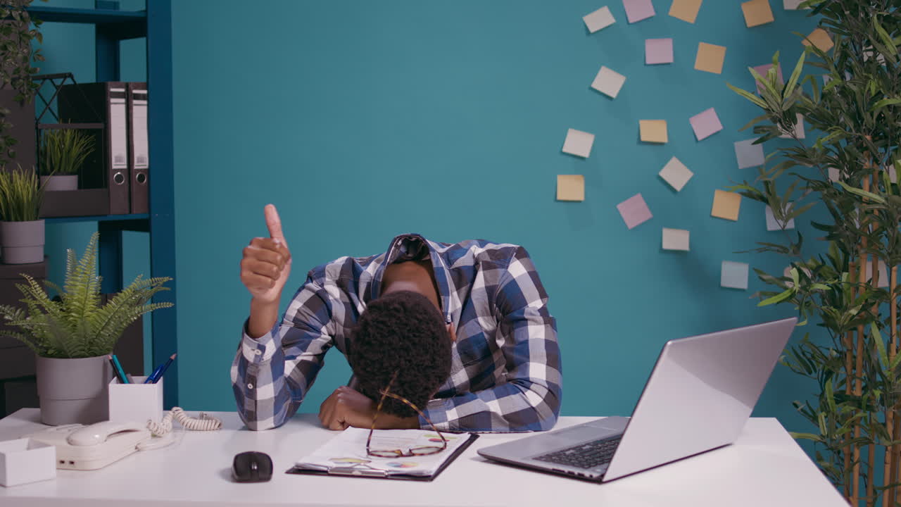 Tired employee resting head on desk and giving thumbs up