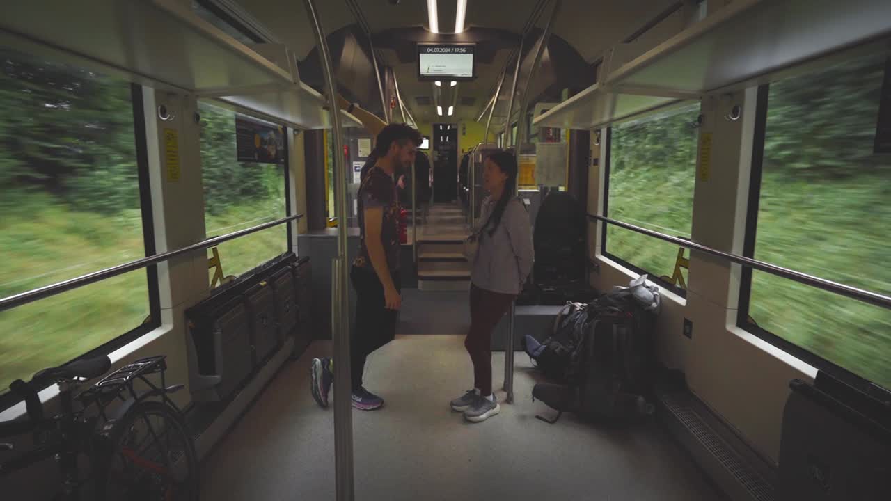 두 명의 승객이 서서 이동하는 열차에서 대화하고, 그들의 여행 장비와 자전거에 둘러싸여, 창문으로 보이는 울창한 초록색으로, 중간 정적
