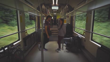 dos pasajeros de pie y charlar a bordo de un tren en movimiento, rodeados por su equipo de viaje y bicicletas, con vegetación exuberante visible a través de las ventanas, estática media