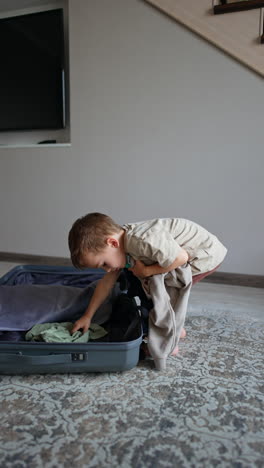 little boy takes his clothes out of suitcase and tosses them up. kid watches his things fall and jumps happily. vertical video.