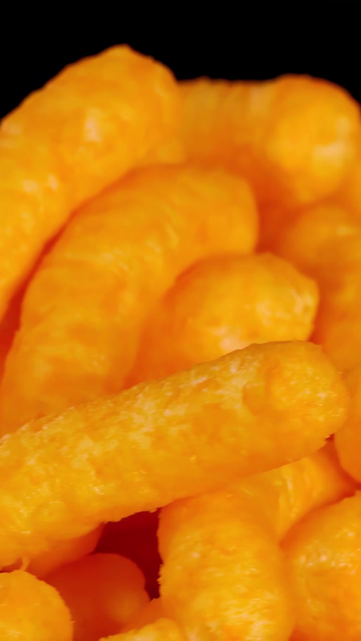 Crispy cheese puff snacks rotate in close-up, vibrant lighting, black background, macro detail