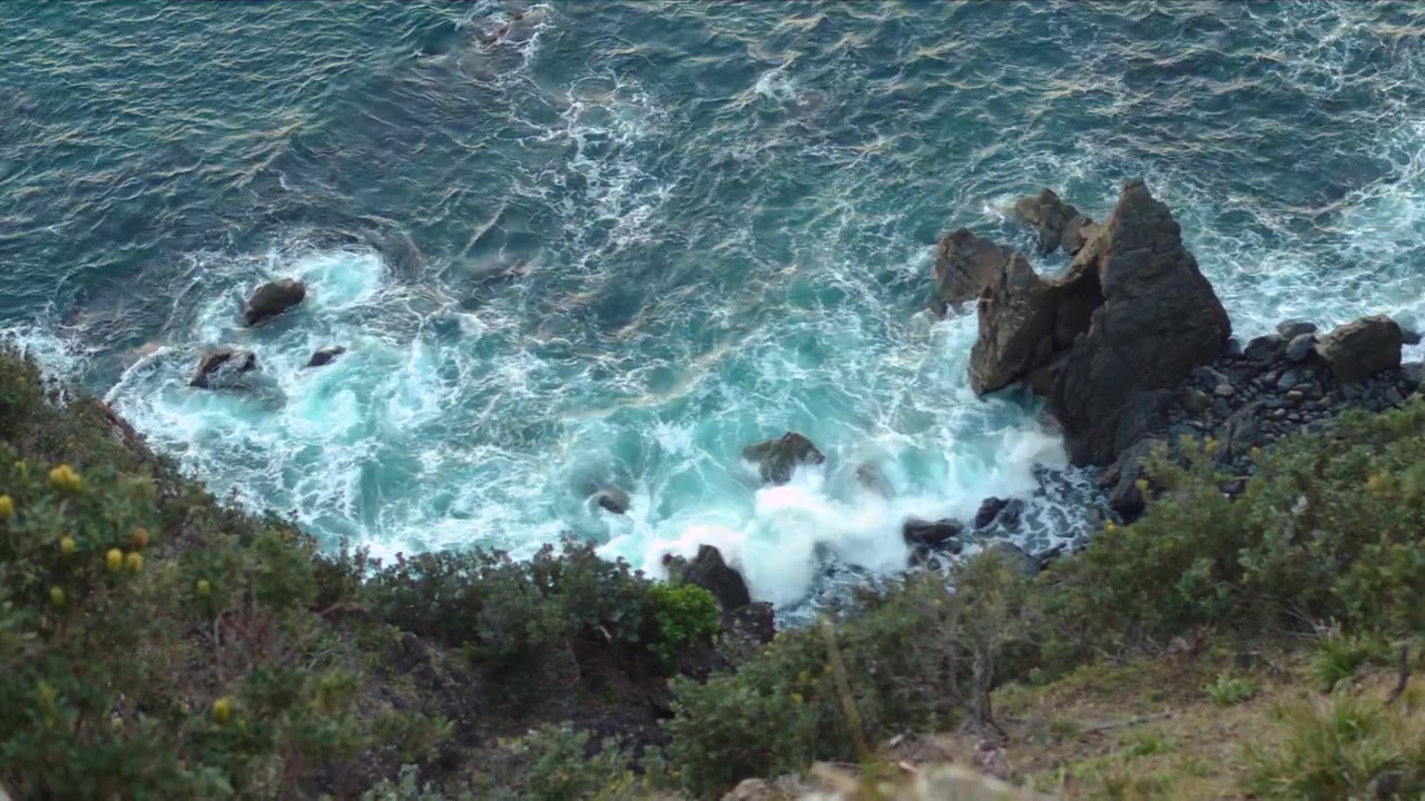 Vista panoramica anamorfica in 4K di onde oceaniche turchesi che si infrangono contro la frastagliata costa rocciosa a Byron Bay, Australia, circondata da vegetazione costiera.