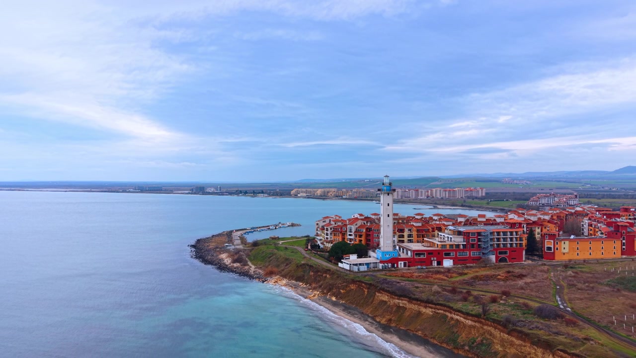 Veduta con drone di una penisola costiera con faro e città turistica nella baia. Un faro bianco si erge sul bordo della scogliera mentre colorati appartamenti del resort si estendono dietro di esso sopra una baia curva color verde acqua
