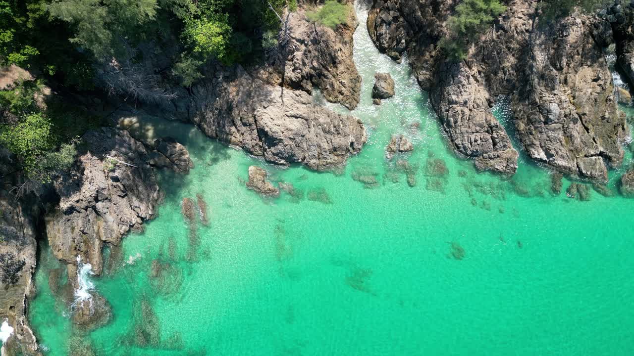 Top-down view of tropical Thai coast with rugged rocks, lush greenery, and crystal-clear turquoise water.
