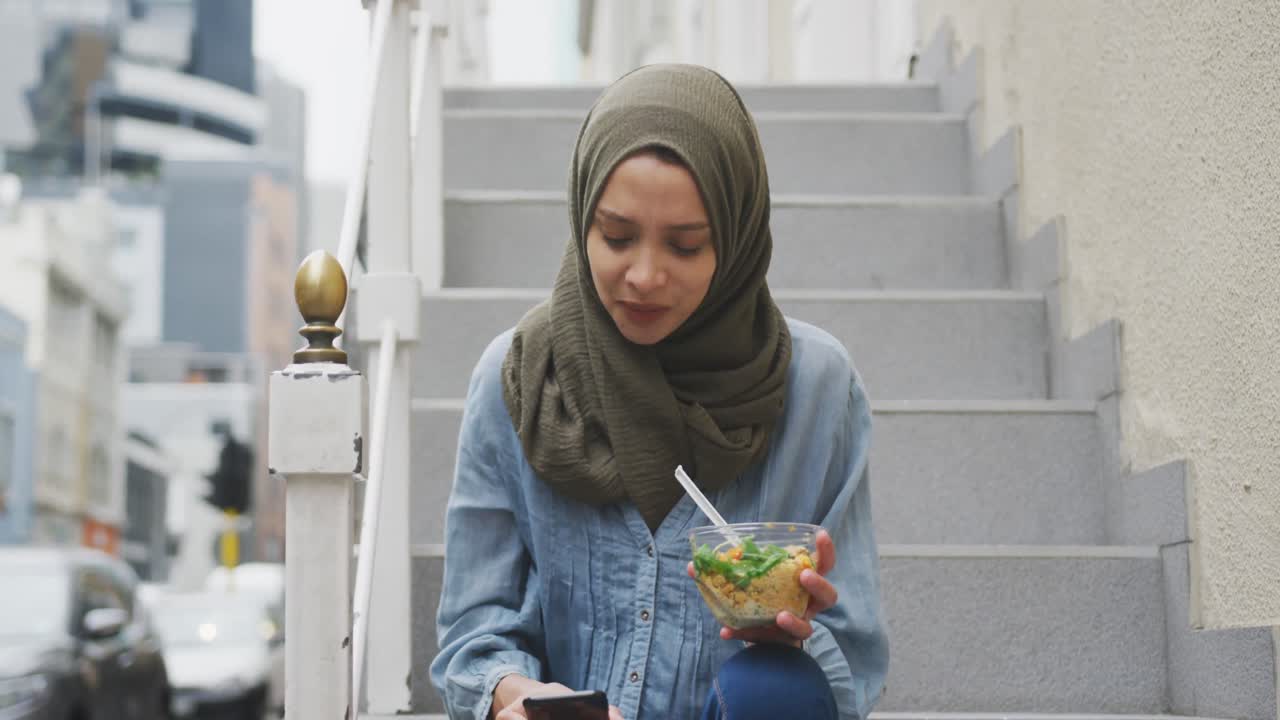 mujer con hijab con una ensalada para llevar usando su teléfono sentada en las escaleras
