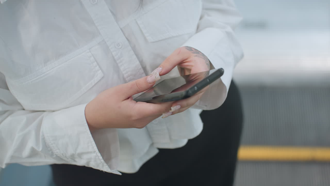 Close up view of person holding smartphone with both hands, focused on screen while stepping off moving escalator in bright indoor shopping environment, showcasing technology use during transit