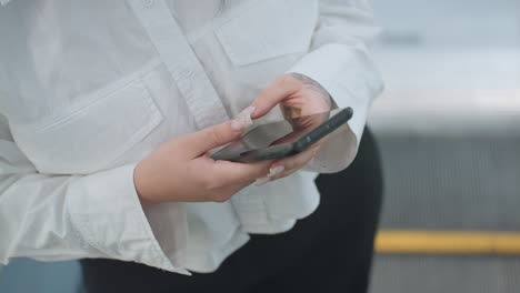 close up view of person holding smartphone with both hands, focused on screen while stepping off moving escalator in bright indoor shopping environment, showcasing technology use during transit