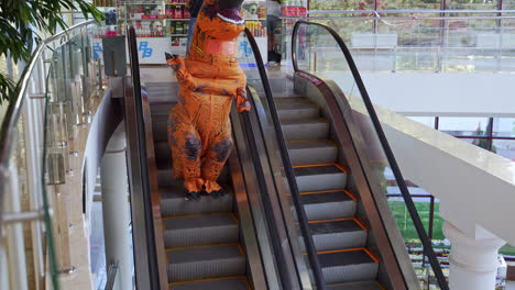 man wearing orange costume of dinosaur goes down by the escalator. spotty t-rex dancing funnily on the stairs. view from top.