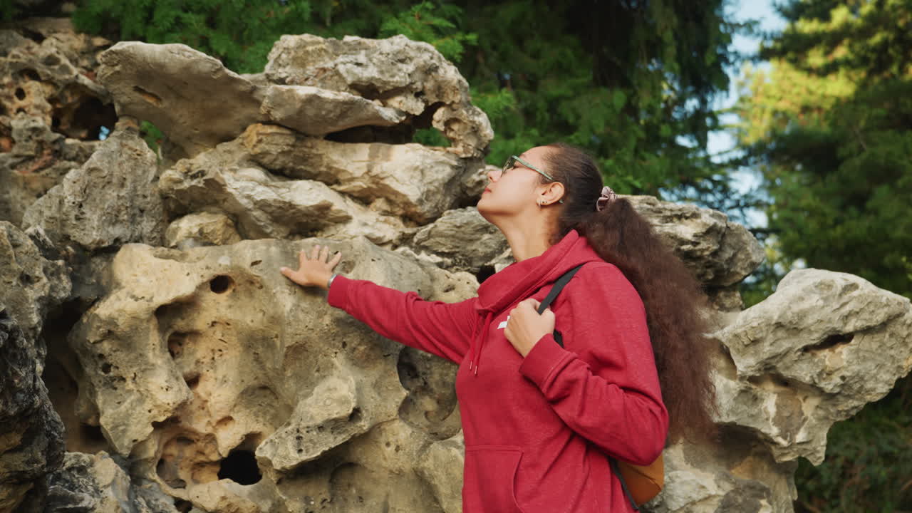 Black woman examines rock textures closely, porous limestone studied with fingertips while leaning against formation, red jacket and yellow backpack visible, curly hair swept back, closeup detail