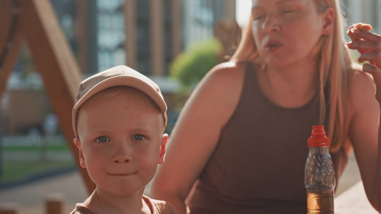 Una donna e un bambino sono seduti insieme all'aperto, il bambino indossa un berretto. L'adulto tiene del cibo mentre entrambi sono vicino a una bottiglia di plastica con tappo rosso. La luce del sole crea tonalità calde in un ambiente di parco urbano.