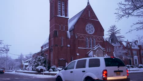 slow pan down of a large brick church in a quiet nj town while it snows with cars passing by