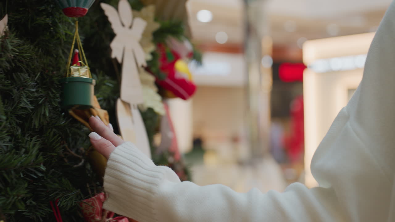 Close-up of lady in white sweater touching decorative Christmas tree affectionately in a shopping area, evoking a warm, festive atmosphere with soft holiday lights in the background