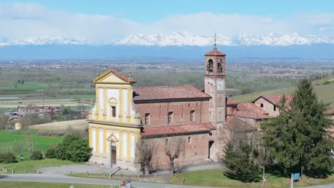 Church-of-Saint-Peter-Apostle-Catholic-church,-Gabiano,-Italy,-Piedmont-region-with-snow-covered-white-peaks-of-the-northern-mountains-before-Switzerland