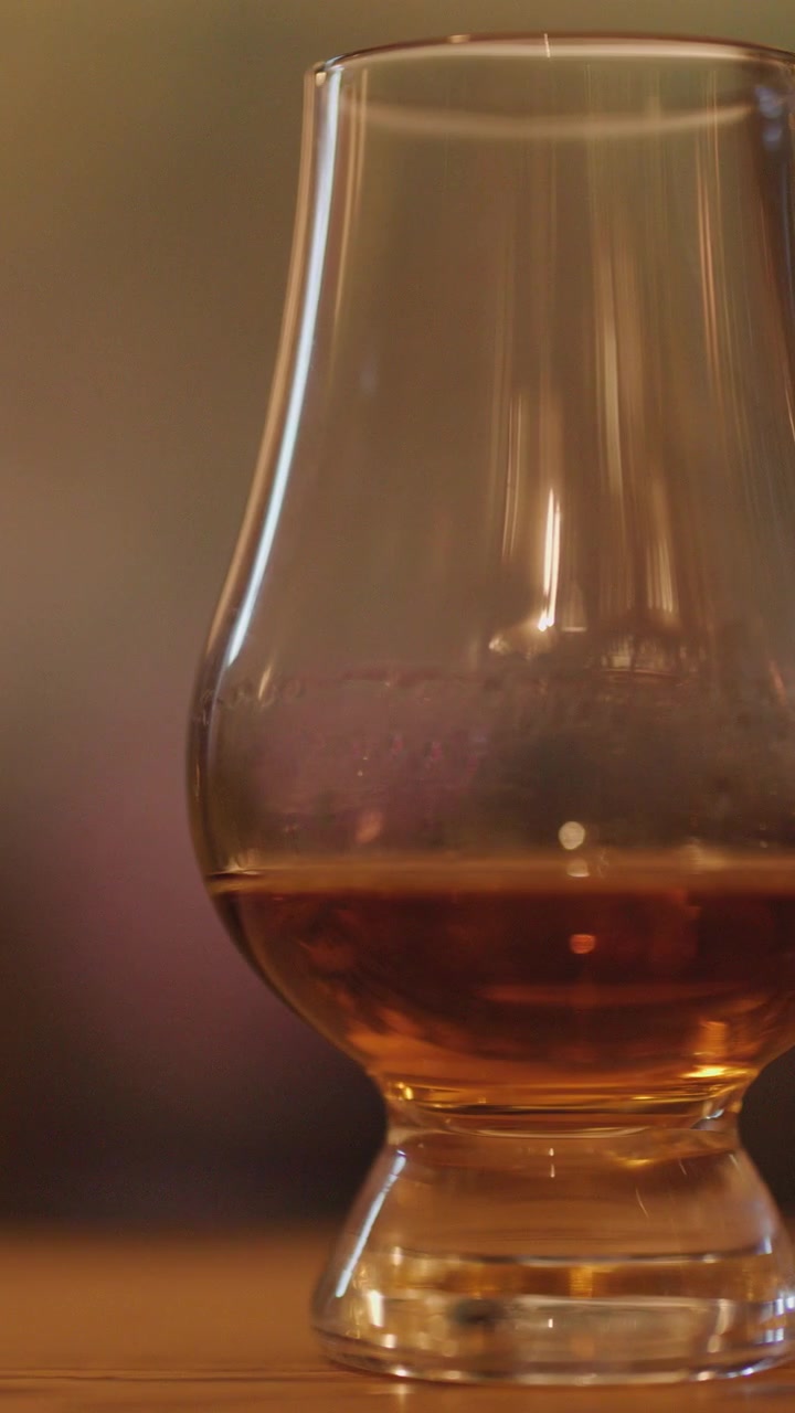 Una fluida panoramica della telecamera rivela bicchieri di whisky con liquido ambrato su un bancone di legno, ambientati su uno sfondo caldamente illuminato e leggermente sfocato in condizioni di scarsa illuminazione