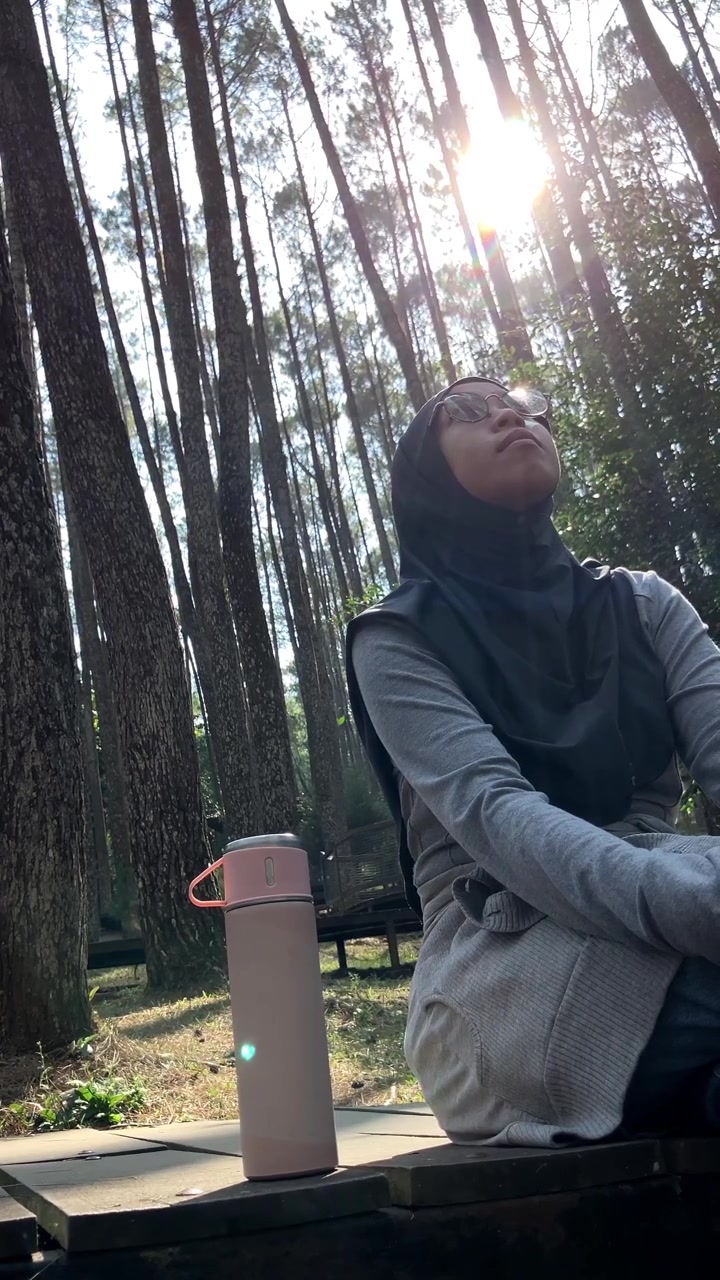 Gerak lambat seorang wanita Muslim muda berhijab duduk dengan termosnya. Dia bersantai dan menikmati pemandangan indah di hutan pinus yang tenang dan cerah. Sempurna untuk konsep gaya hidup alam luar ruangan.