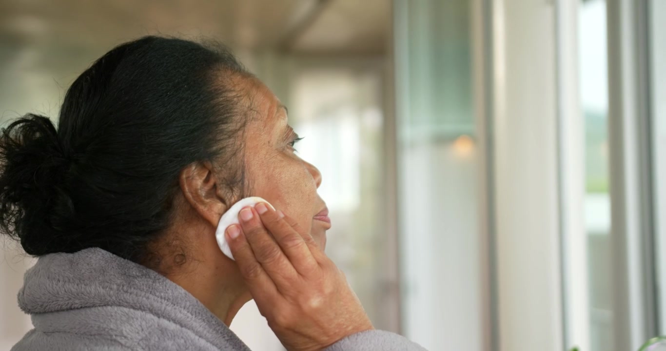 Senior African American woman in gray robe standing at sink by window raising pad removing makeup