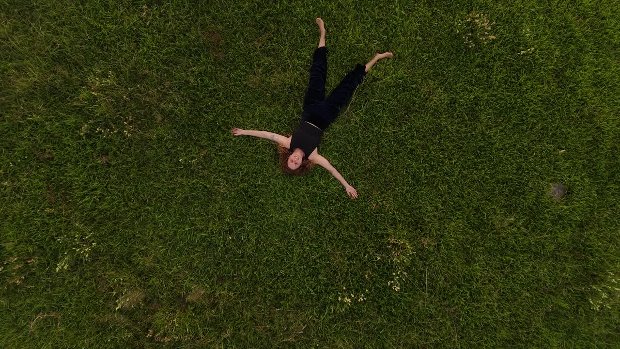 Barefoot woman lying on grass field and looking up to the sky. Aerial top down rising shot. Brunette girl relax in nature.