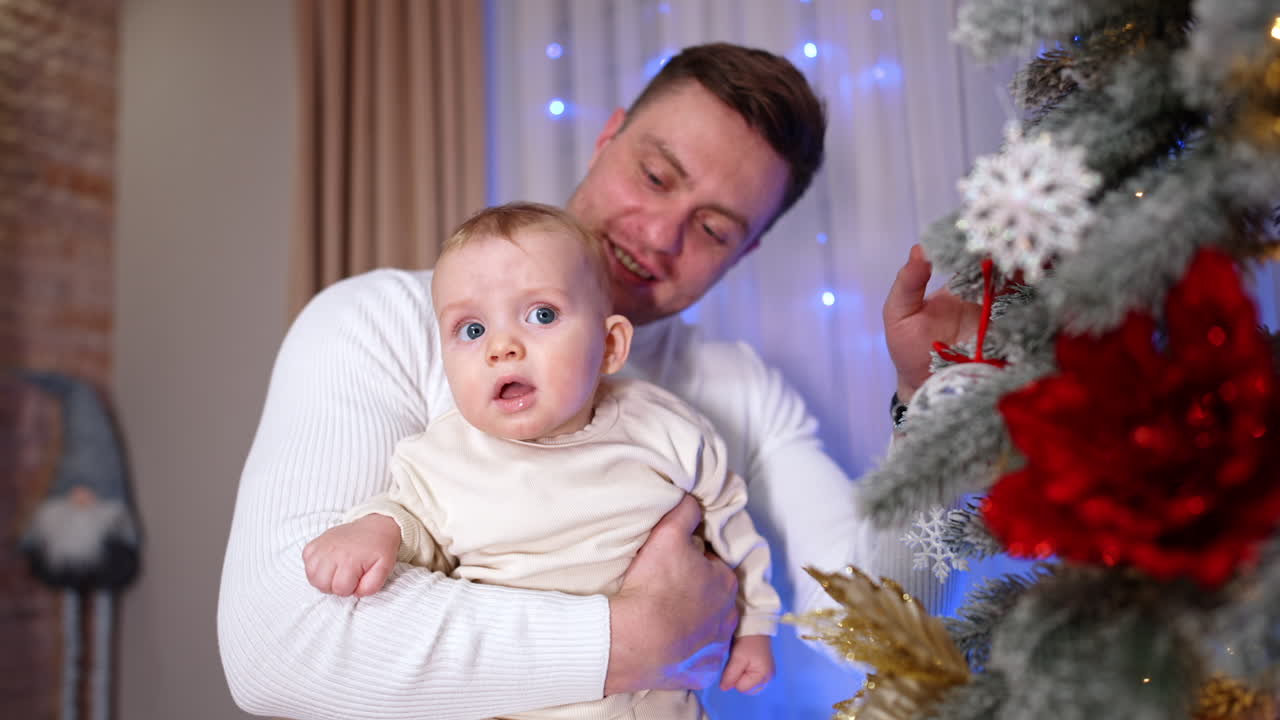 Father is holding his adorable baby standing near the Christmas tree. Little child looks surprised and astonished.