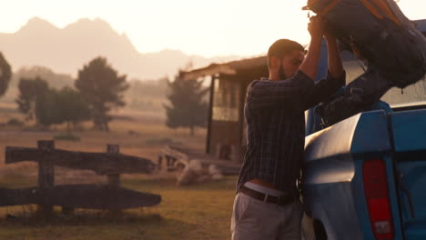 portrait of man unloading backpacks from pick up truck on road trip to cabin in countryside