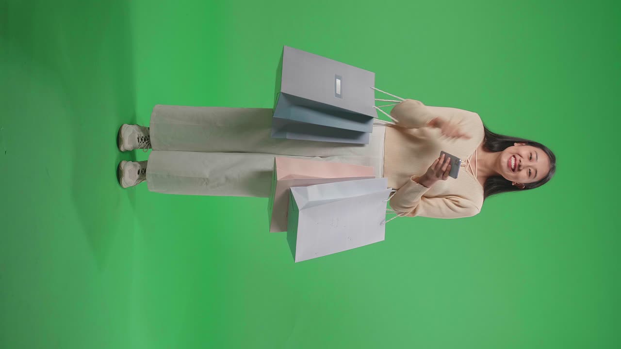 vista completa de una mujer asiática de compras con bolsas de compras siendo feliz ser capaz de usar el teléfono móvil de compras en línea mientras está de pie frente a la pantalla verde