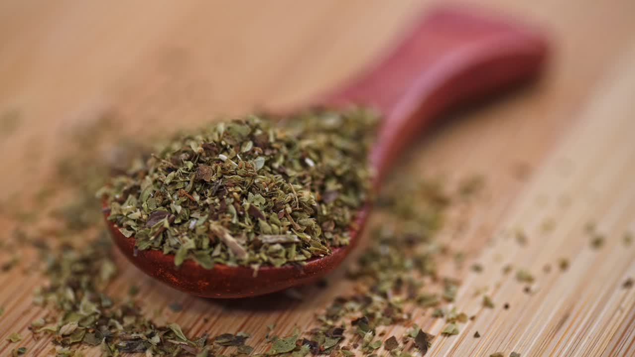 A close-up of dried oregano on a wooden spoon, placed on a wooden surface. The finely chopped herb is slightly spilling over, showcasing its rich green and brown tones.