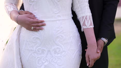 a beautiful bride stands in a white dress, the groom stands beside her, his hand resting gently on her belly in an intimate gesture of love and support