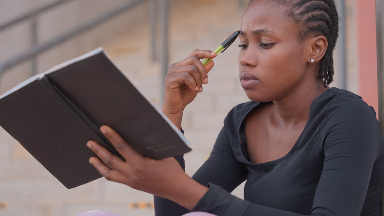 Scrittrice di colore che sfoglia pagine con una penna sulle scale, in una pausa pensierosa, mentre controlla gli appunti e modifica il diario, sullo sfondo di un campus informale e con luce naturale. Primi piani mostrano pianificazione e decisioni.