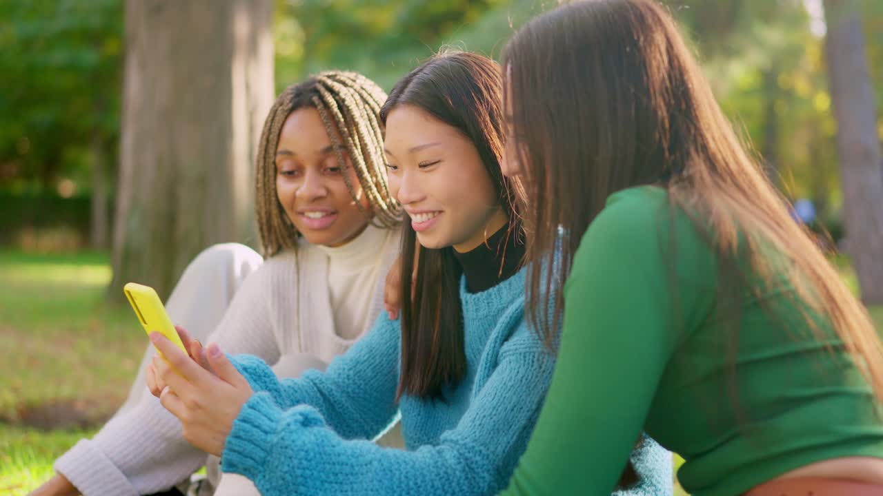 Three young women friends sharing a smartphone outdoors in a park