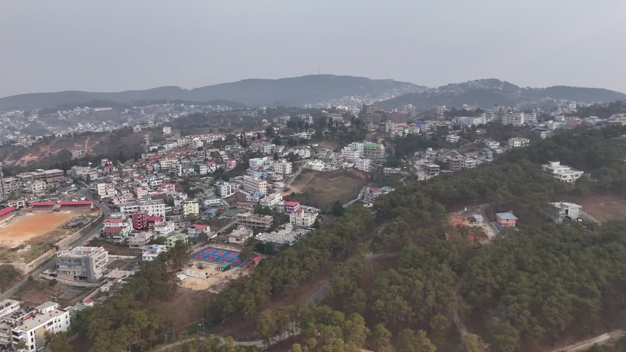 Riprese con drone rivelano una vasta città collinare con edifici a più piani colorati immersi tra lussureggianti foreste di pini verdi. Montagne si ergono sullo sfondo nebbioso mentre strade tortuose attraversano il terreno.