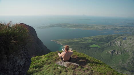 excursionista sin camisa sentado en una cornisa rocosa tomando fotos de viaje usando una cámara de lente larga en el monte donnamannen, nordland, noruega