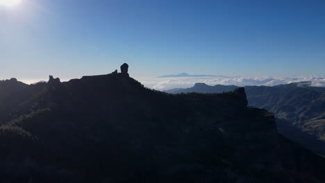 Flying-through-the-air-in-the-midst-of-treetops-toward-the-famous-big-rock,-Roque-Nublo,-in-Gran-Canaria-during-a-beautiful-blue-sky