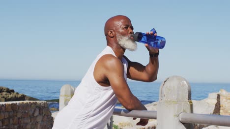 Senior-african-american-man-exercising-drinking-water-on-rocks-by-the-sea