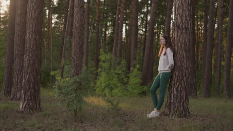 lonely woman with headsets rests in forest. young lady listens to calm music enjoying autumn sunset leaning on tree. relaxation moment in wild nature