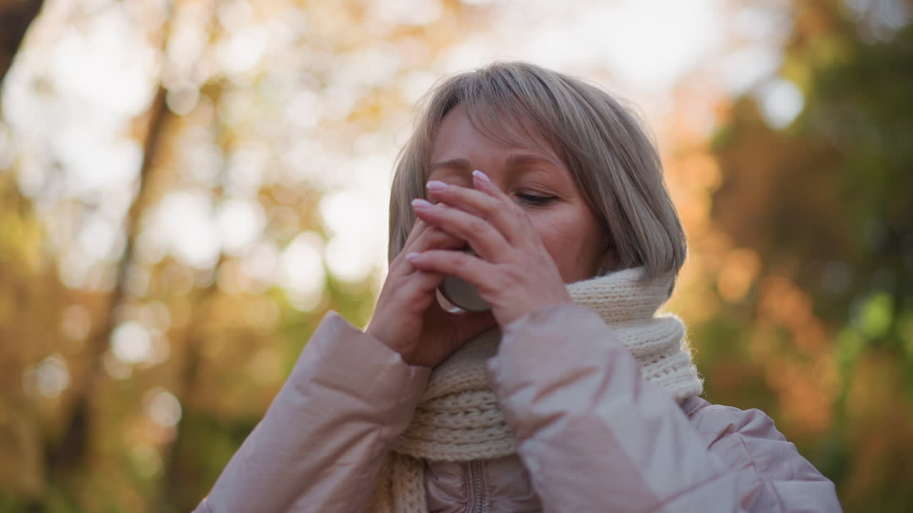 close up of woman holding cup of hot tea near lips, blowing to cool steaming drink, wrapped in knit scarf as sunlight glow frames face against blurred autumn foliage
